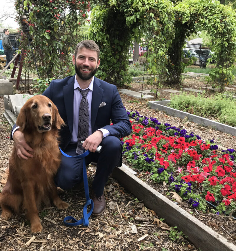 Thrive Recovery founder Sammy in a suit kneeling down with his golden retriever next to a flower bed